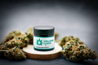 Jar of Wellness by Wes Premium Hemp Extract salve positioned against a backdrop of dried hemp buds on a wooden surface, highlighting its natural ingredients.